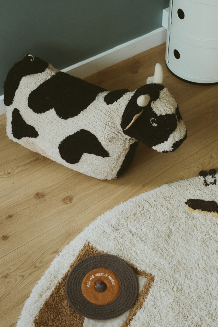 Plüschiges, kuhförmiges Bodenkissen in Schwarz und Weiß, daneben ein runder Teppich mit Plattenspielermotiv auf Holzboden.
