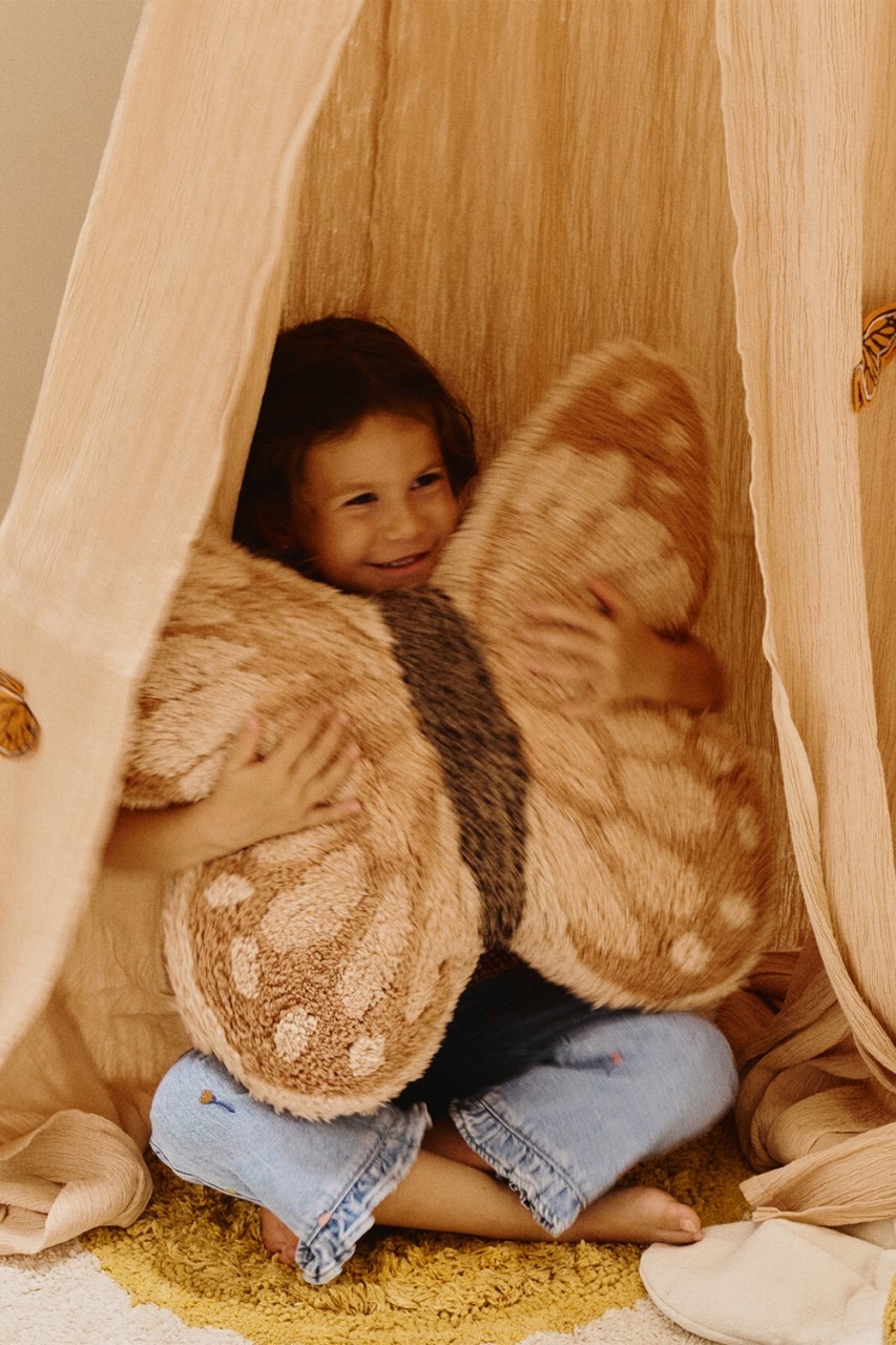 Ein kleines Kind sitzt in einem beigen Stoff-Tipi und umarmt ein großes, braunes Plüschkissen in Form eines Schmetterlings.