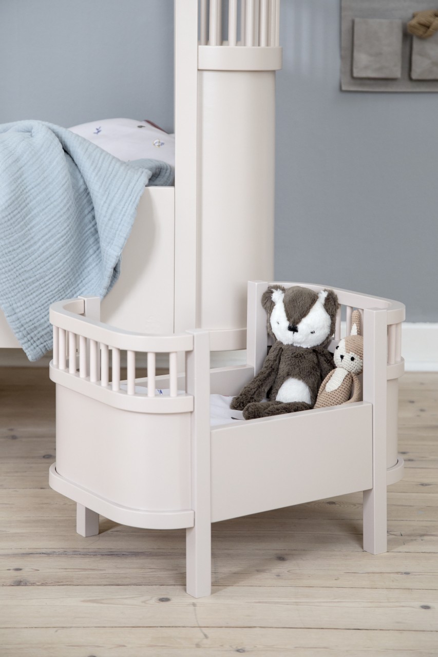 Hellrosa Puppenbett aus Holz mit einem Plüschdachs und einem kleinen gehäkelten Hasenspielzeug darin auf einer weißen Matratze.